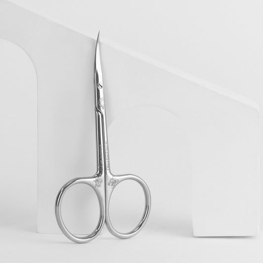 A pair of Staleks PRO Professional cuticle scissors (magnolia) EXCLUSIVE 20 TYPE 2 with pointed tips, designed for a precise cuticle cut, resting against a white, curved stand with a minimalist design.