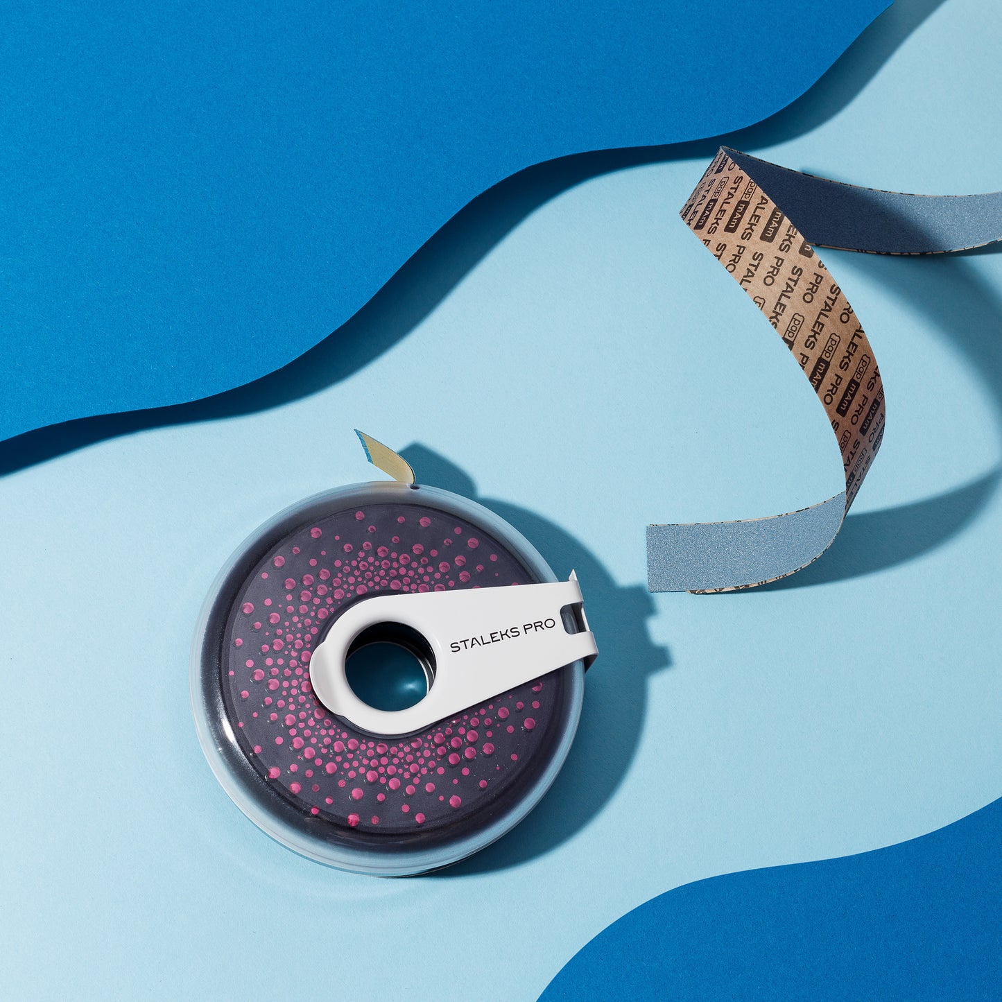 A pink and black Staleks pro tape dispenser with a clear center, resting on a blue background with wavy paper layers. A piece of STALEKS PRO Nail File Refill Roll White Disposable Abrasive Tape papMam EXPERT in a Plastic Case ATC with measurements is partially pulled out.