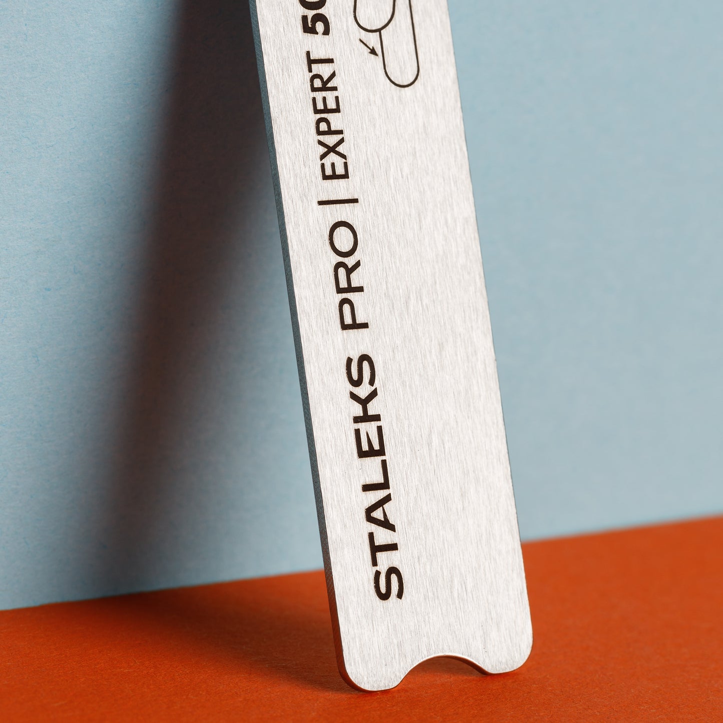 Close-up view of a stainless steel nail file with "Staleks EXPERT 50" engraved on it, set against a dual blue and orange background.
