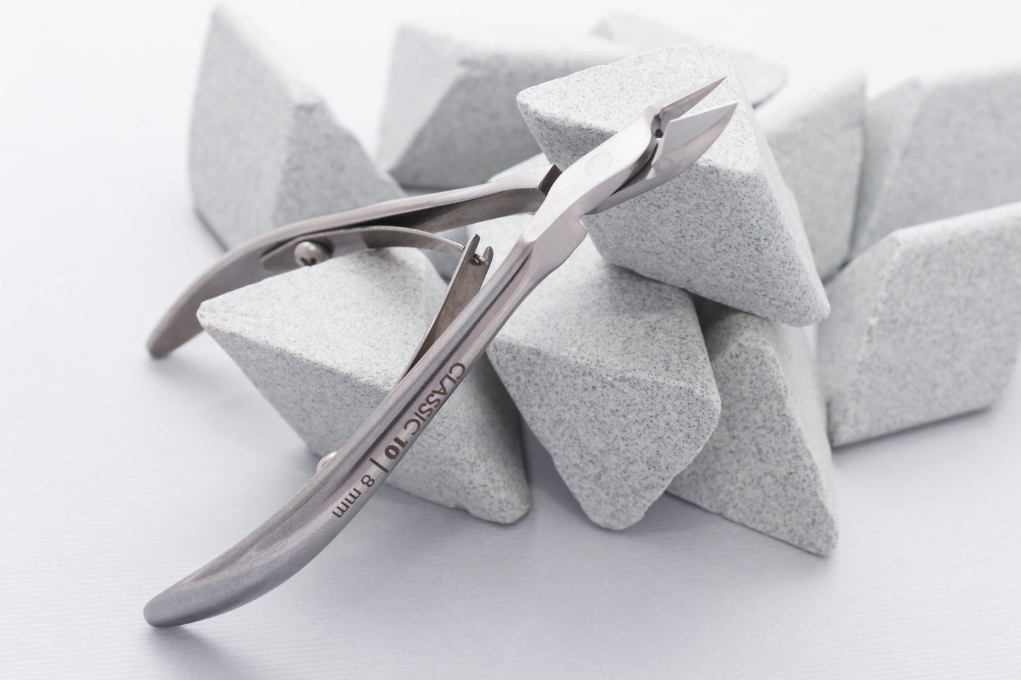 A pair of Staleks stainless steel, diamond sharpened cuticle nippers 8 mm jaw CLASSIC 10 rests atop several porous grey pumice stones on a white background. The tool is angled, displaying its sharp, precise blades.