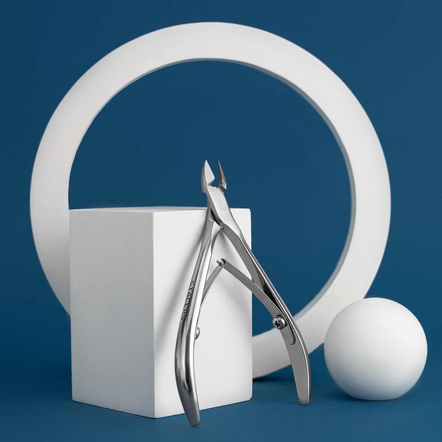A minimalist still life featuring a pair of Staleks Cuticle nippers 8 mm jaw CLASSIC 10 standing on a white cube with a white sphere and a large circular white ring against a solid blue background.