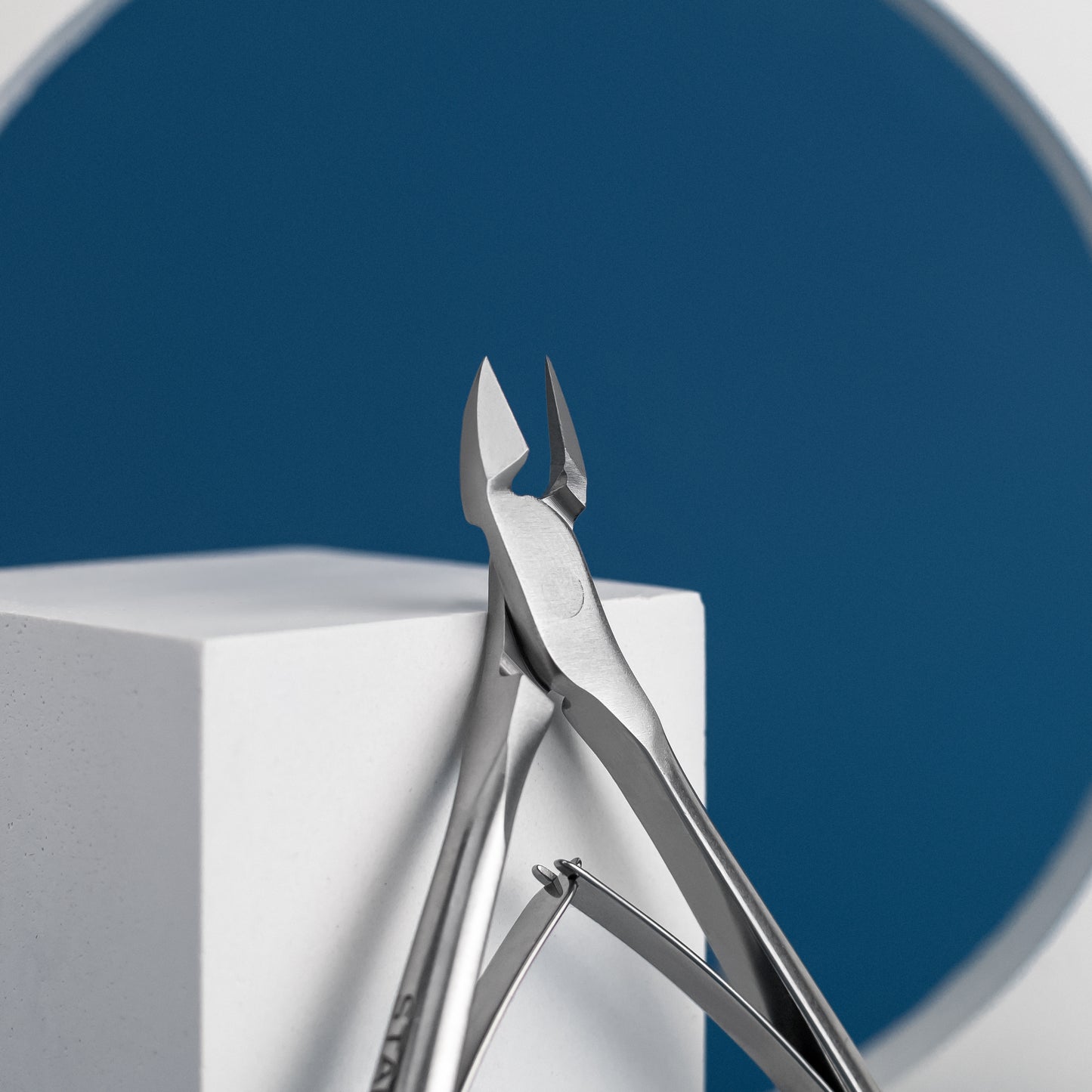 Close-up of Staleks PRO Cuticle nippers 8 mm jaw CLASSIC 10 by Staleks resting on a white geometric object with a circular blue backdrop. The focus is on the precise, diamond-sharpened edges of the nippers' cutting.