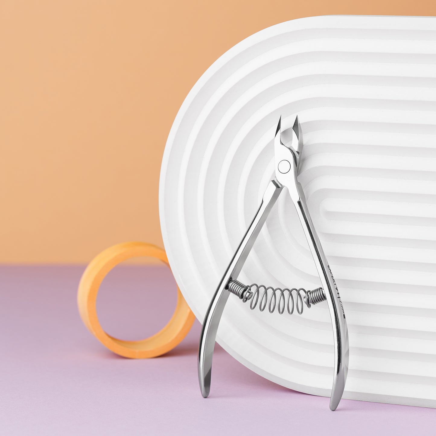A Staleks STALEKS PRO Professional cuticle nipper 7 mm jaw EXPERT 91 rests in front of a large white spiral decoration with orange round rings, set against a dual-colored orange and purple background.
