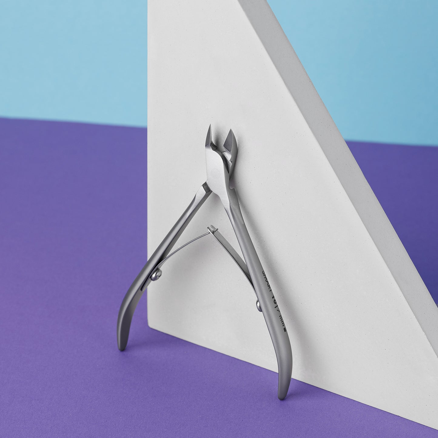 A pair of Staleks STALEKS PRO Professional cuticle nippers 7 mm jaw SMART 10 leaning against a white triangular block on a purple and blue background.