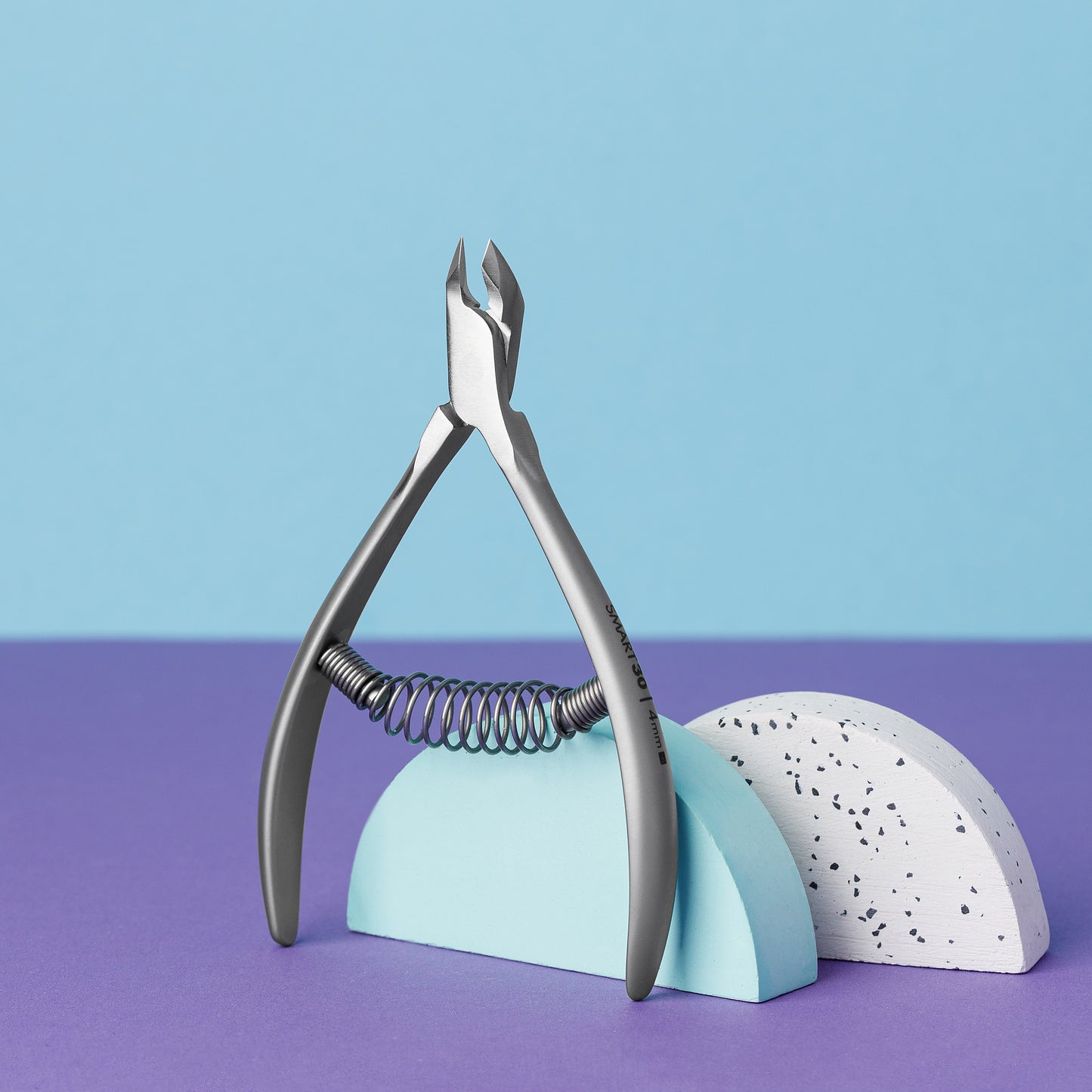 The STALEKS PRO Professional Cuticle Nippers 4 mm jaw SMART 30, made of stainless steel with a spring mechanism, rest elegantly on two half-cylinder objects—one light blue and one white with black speckles. The split blue and purple background highlights the nippers' sleek Staleks design.
