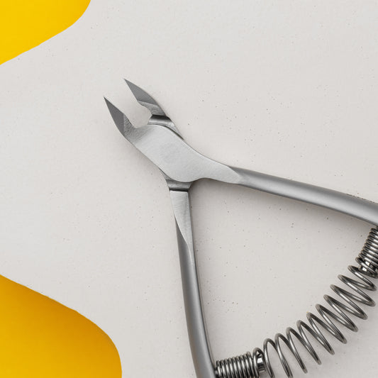 A close-up of Staleks stainless steel nail clippers with a spiral spring, laid diagonally across a background with white and yellow sections.
