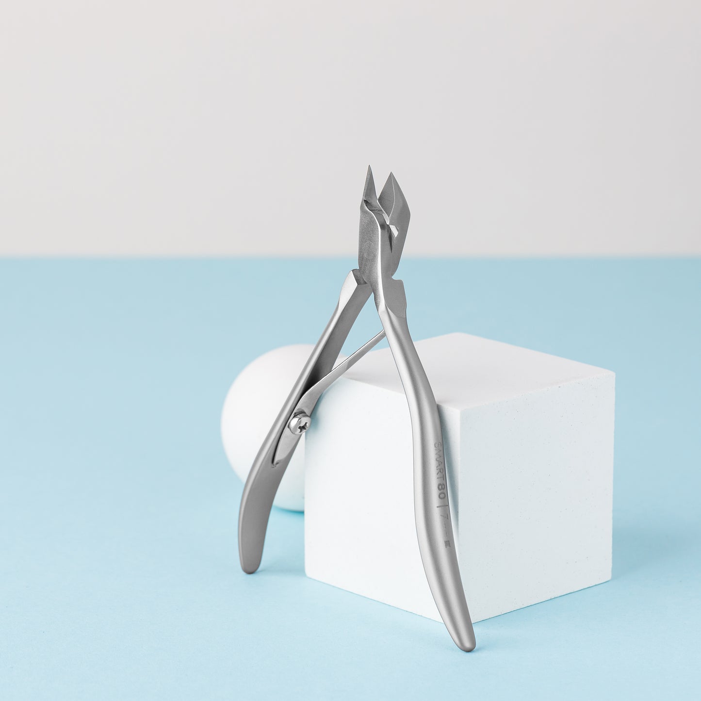 Close-up of STALEKS PRO Professional cuticle nippers SMART 80 7 mm standing open on a white cube against a minimalist background. The setting includes a light blue surface and a white wall, enhancing the clean and simple aesthetic ideal for any professional manicure or pedicure session.