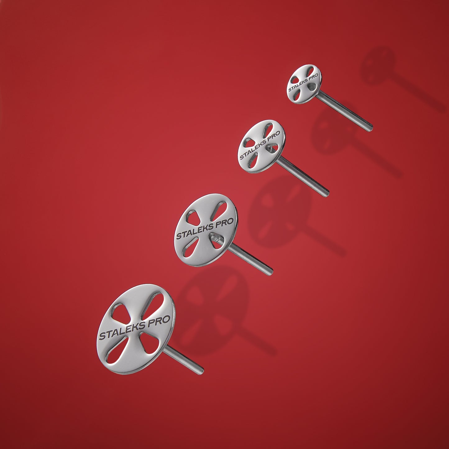 Five circular, silver-colored stainless steel STALEKS PRO pedicure tool pins with "Staleks" branding, arranged and scattered on a vibrant red background, casting soft shadows.