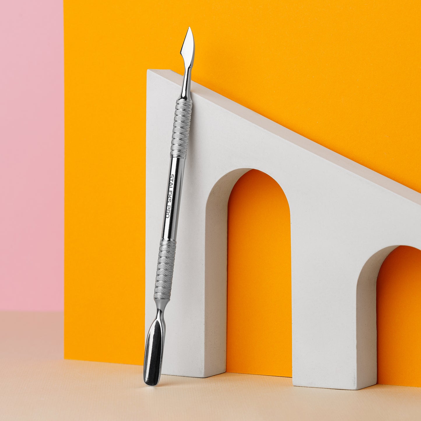 A Staleks STALEKS PRO Manicure pusher EXPERT 30 TYPE 3 (rounded wide pusher and cleaner) with a detailed handle crafted from AISI 420 stainless steel rests on a sleek, white arch set against a peach and orange background, highlighting the tool's precision design.