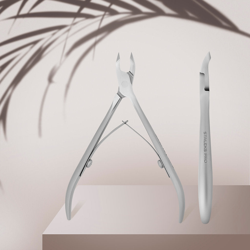 Two STALEKS PRO Professional Cuticle Nippers SMART 10 by Staleks, one open and one closed, are displayed on a light pink platform with soft leafy shadows in the background.