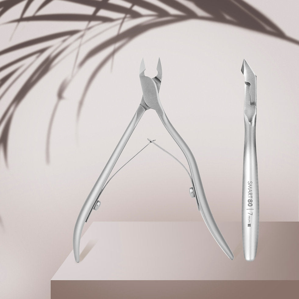 The STALEKS PRO Professional Cuticle Nippers SMART 80 by Staleks and a stainless steel cuticle pusher are displayed on a beige platform with a soft background and leaf shadows.