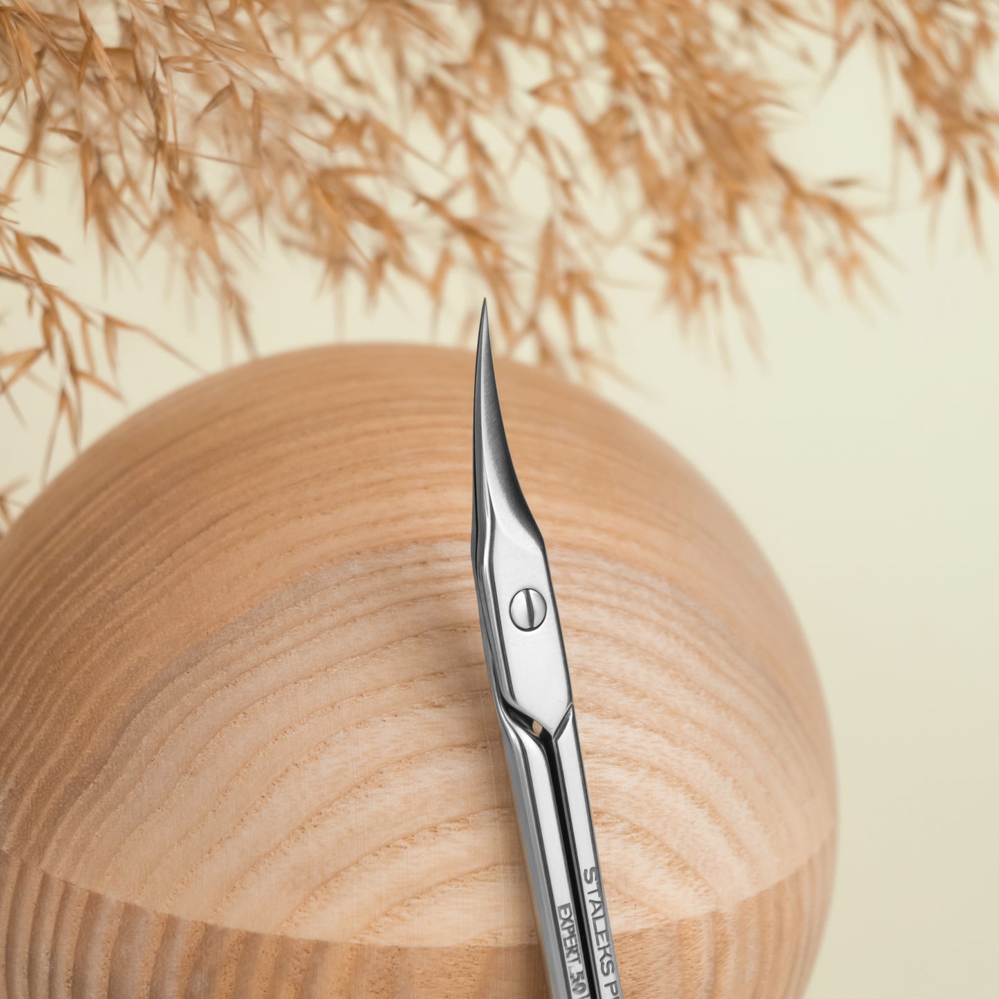 A pair of Staleks STALEKS PRO Professional cuticle scissors EXPERT 50 TYPE 2 with sharp, curved blades designed for nail cuticle cutting rests on a smooth, wooden sphere with dry pampas grass in the background. The setting features a soft