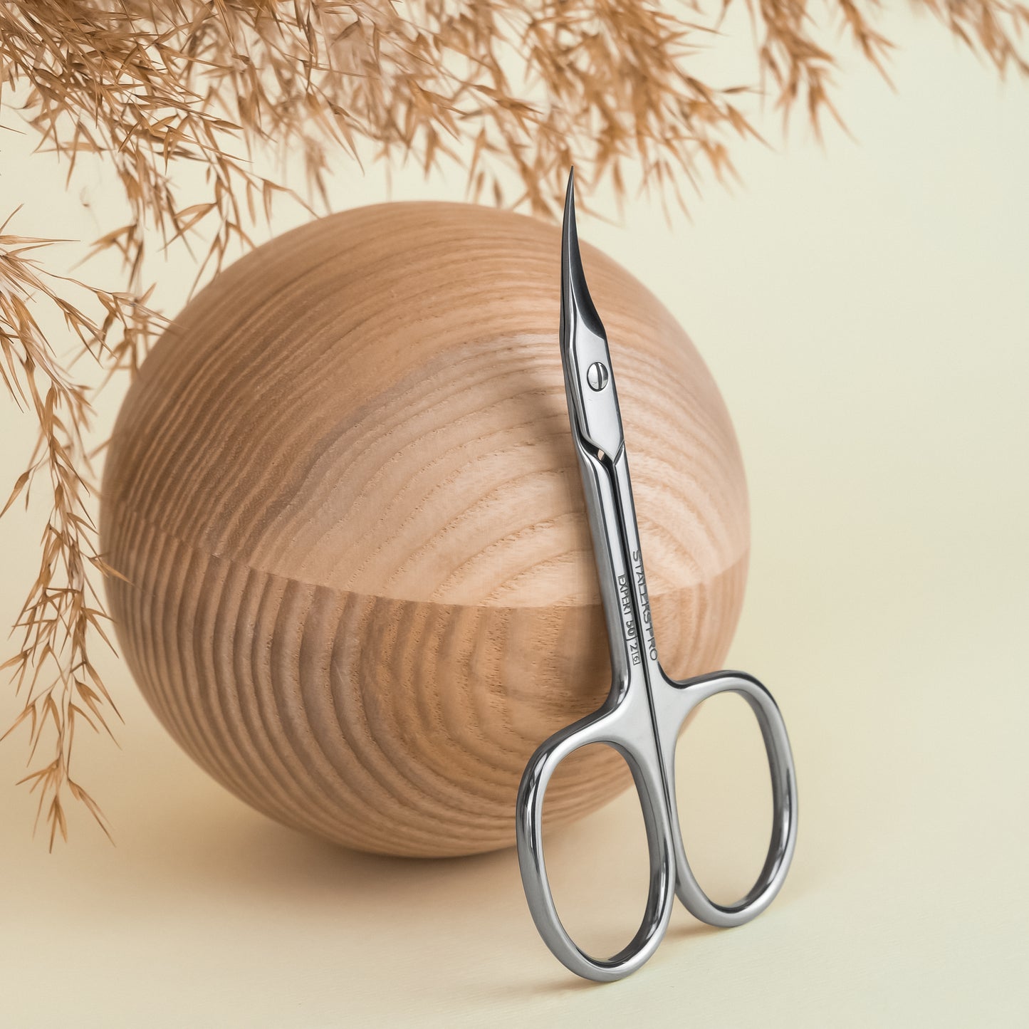 A pair of Staleks Professional cuticle scissors EXPERT 50 TYPE 2 with a blade curve leans against a smooth, spherical wooden object, surrounded by delicate dried plants on a soft beige background.