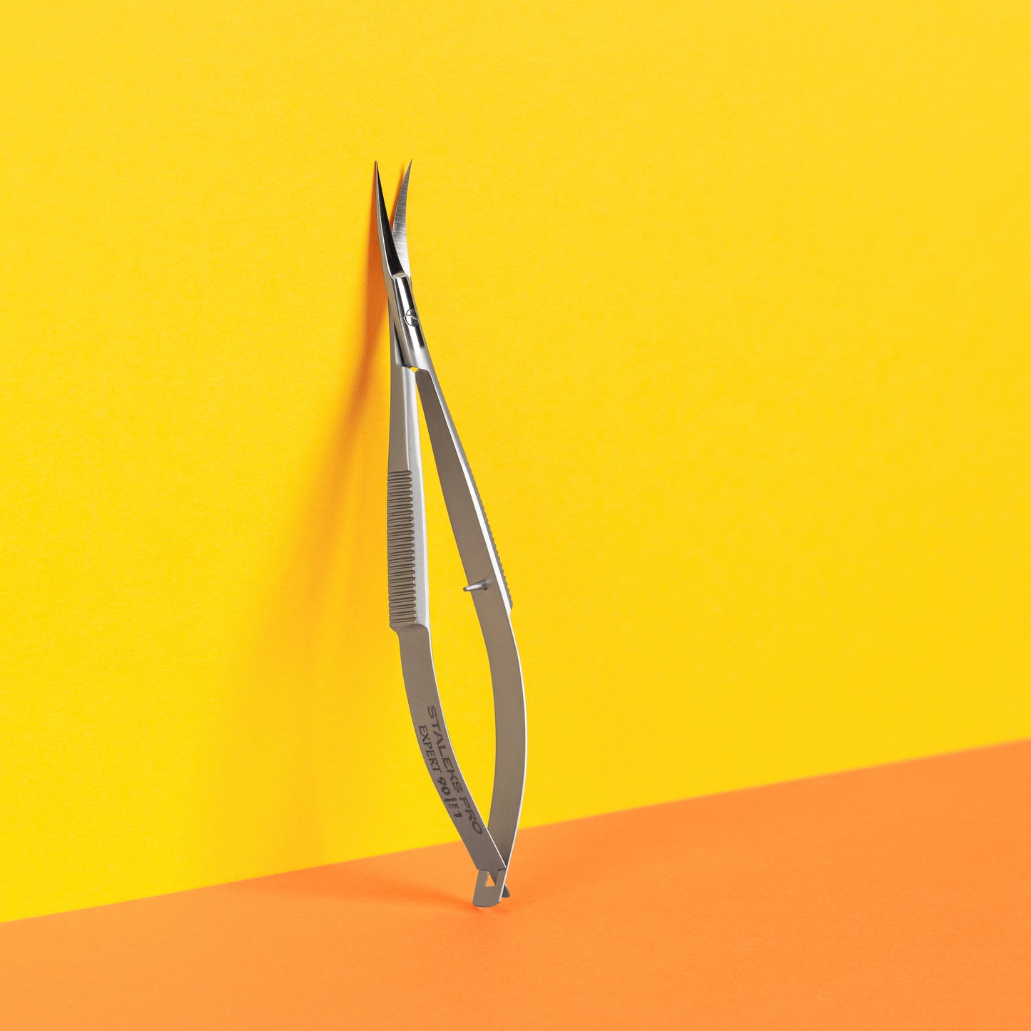 A pair of Staleks STALEKS PRO Professional micro scissors EXPERT 90 TYPE 1 standing upright against a two-tone background, with a yellow top half and an orange bottom half.