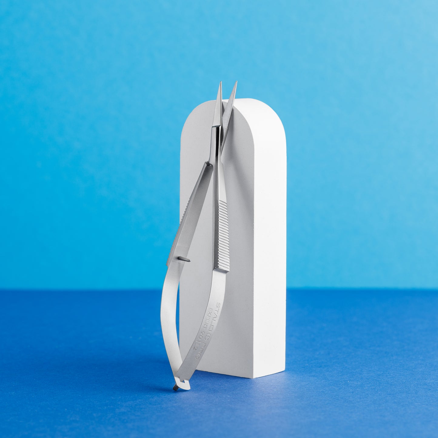 A pair of Staleks Professional micro scissors for eyebrows modeling EXPERT 90 TYPE 2 leaning against a white curved stand, set against a vibrant blue background.