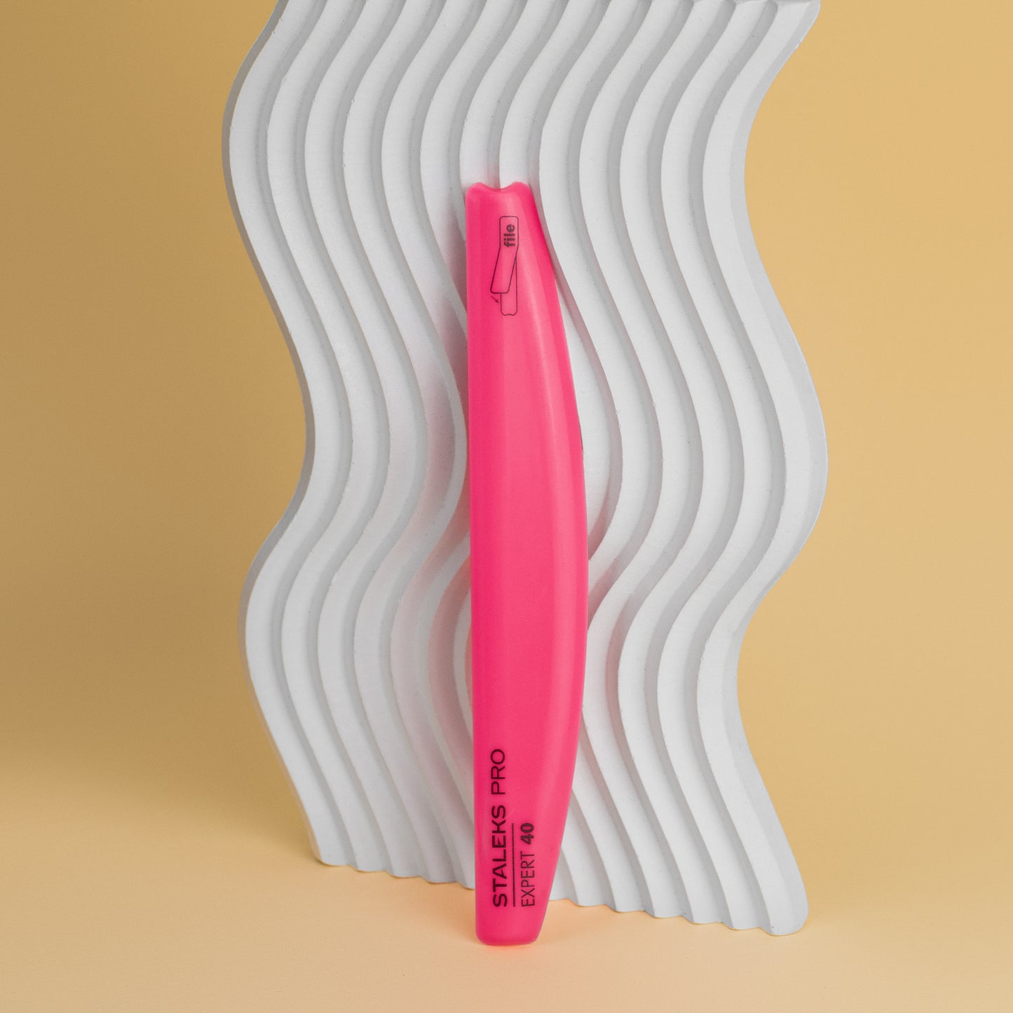 A vibrant pink STALEKS PRO Slanted plastic nail file, crescent (base) EXPERT 40 stands against a white, wavy DFEX-40-150 background on a soft yellow surface, emphasizing texture and color contrast.