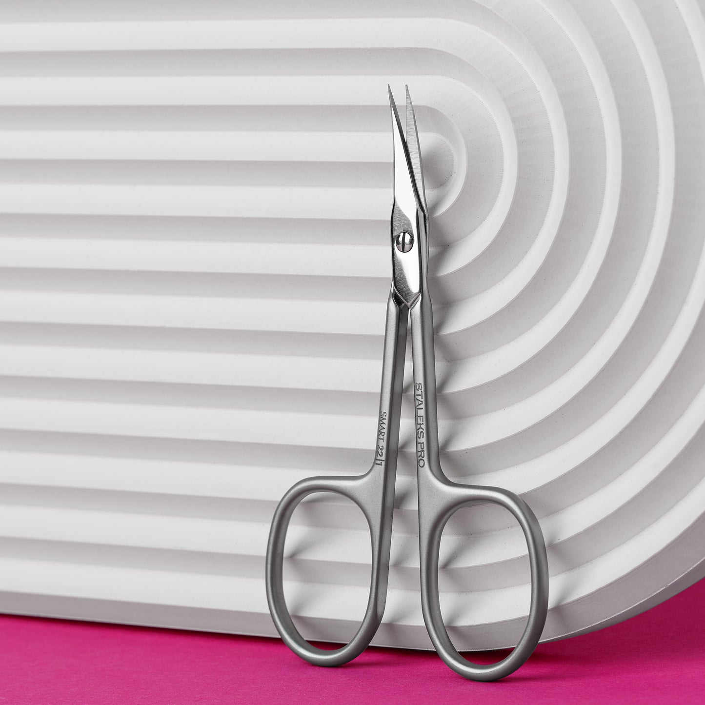 A pair of STALEKS PRO Professional cuticle scissors SMART 22 TYPE 1 by Staleks resting on a white corrugated surface with a vibrant pink background.