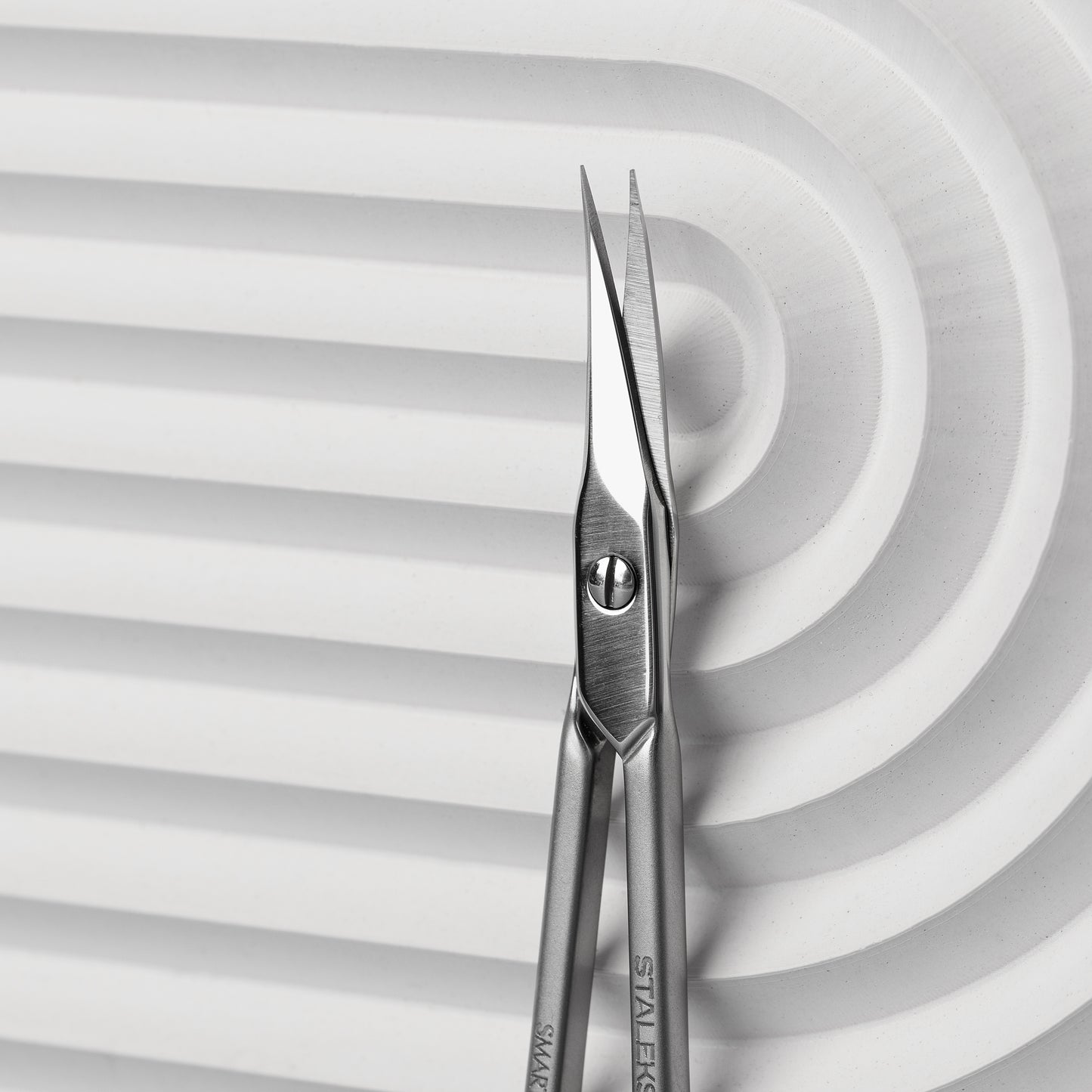 A close-up photo of Staleks Professional cuticle scissors with sharp, pointed tips and narrow straight blades resting on a white, wavy-textured background.