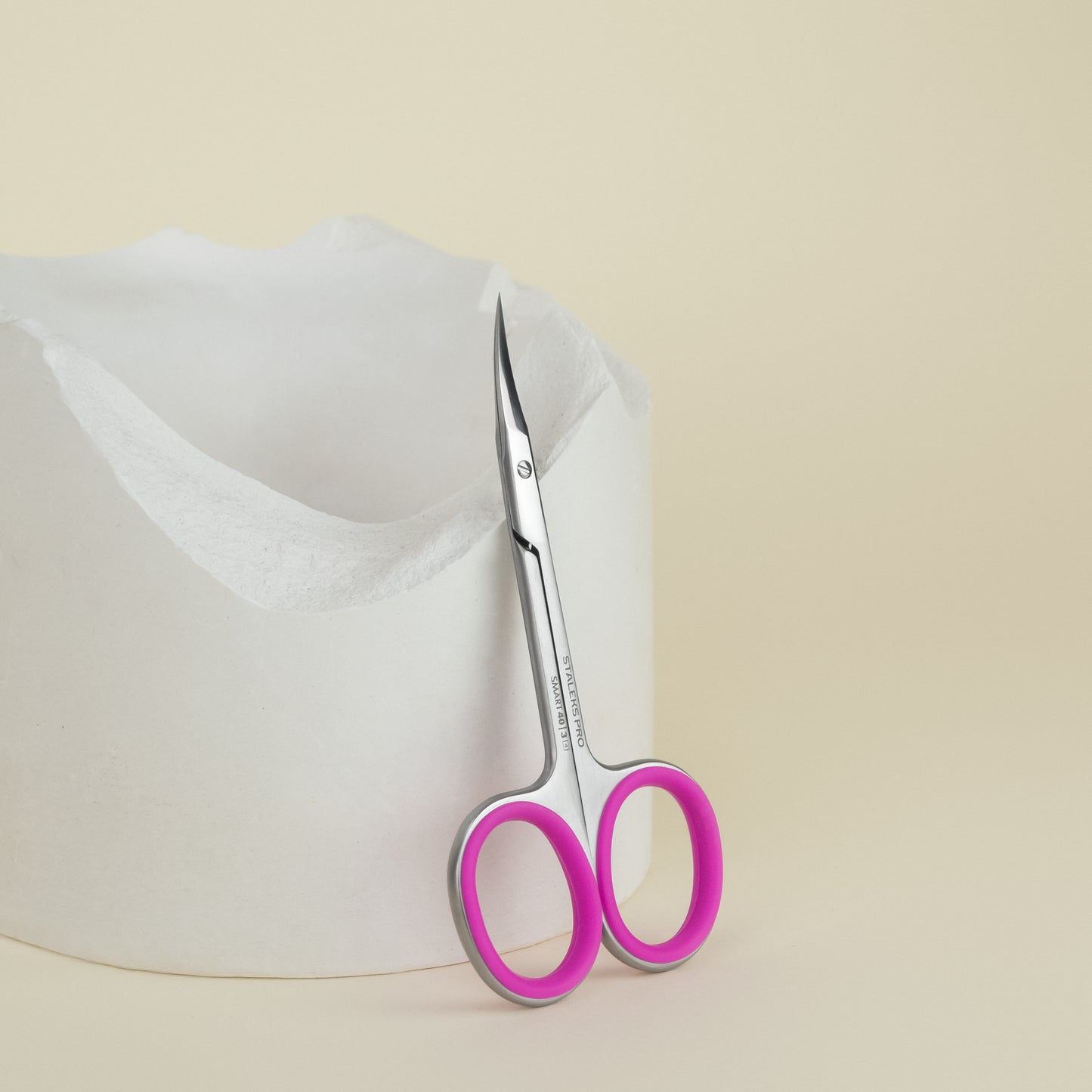 A pair of Staleks Professional cuticle scissors SMART 40 TYPE 3 with bright pink handles poking out of a white paper bag on a beige background.