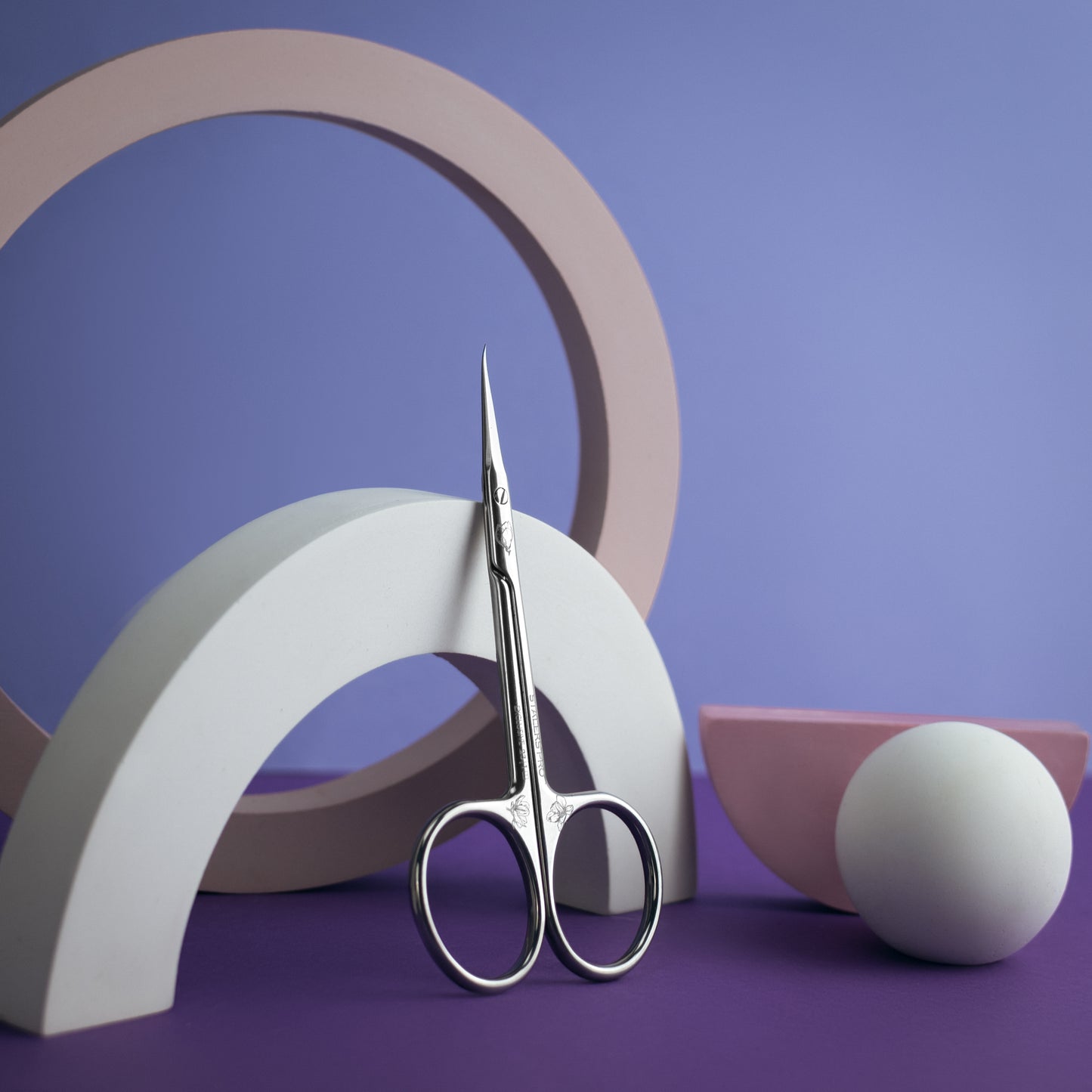 A pair of Staleks Professional cuticle scissors elongated handles (magnolia) TYPE 1 stands upright among geometric shapes in shades of purple and white, creating a minimalist artistic composition.