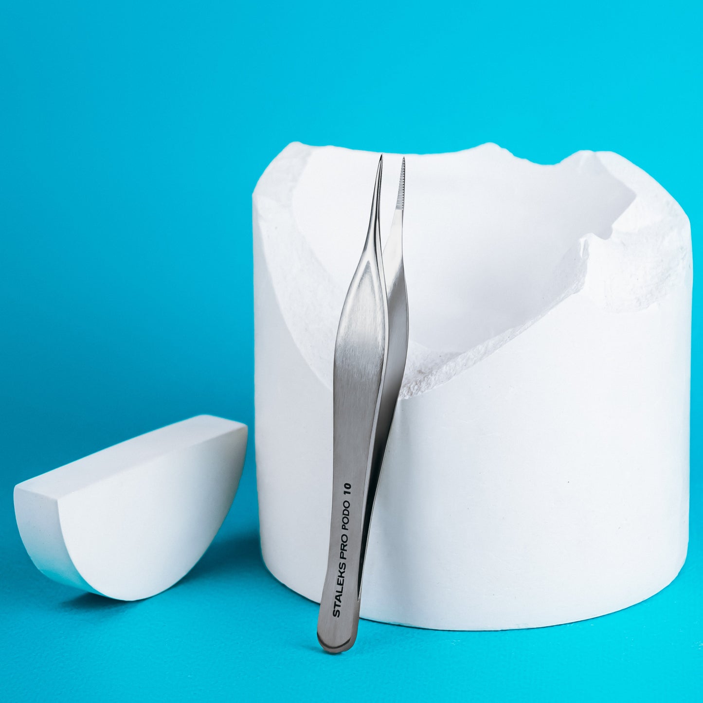 A pair of rust-resistant Staleks STALEKS PRO Podology Splinter Tweezers PODO 10 stands upright against a white cylindrical plaster object with a broken edge on a bright blue background. Perfect for podology splinter or ingrown nail care.