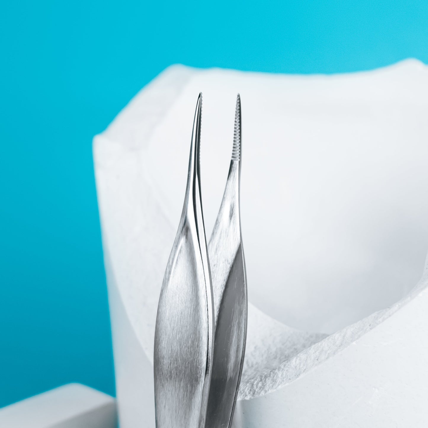 A close-up of two rust-resistant STALEKS PRO Podology Splinter Tweezers PODO 10 by Staleks, with pointed tips, displayed on a bright blue background next to a white dental model—ideal for ingrown nail care or podology tasks.