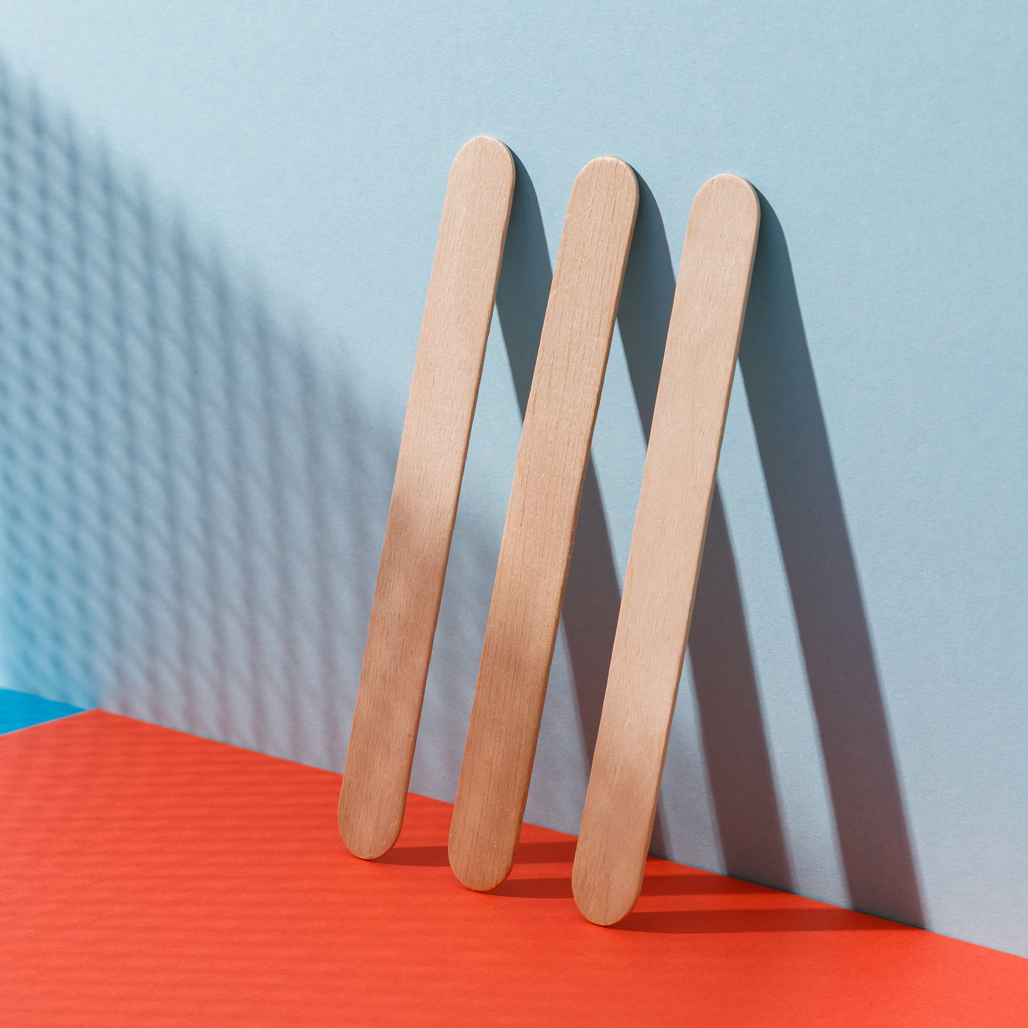 Three STALEKS PRO Disposable wooden Nail files straight (base) EXPERT 50 pcs standing upright and arranged parallel to each other on a surface split between red and blue, with a shadow pattern on a blue background.