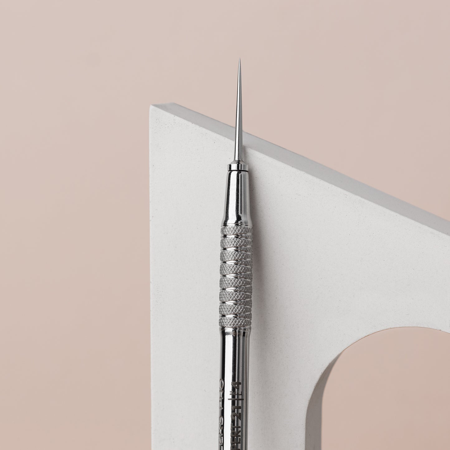 A close-up of a Staleks STALEKS PRO Beauty care double-ended tool (UNO small and Vidal needle straight) EXPERT 21 TYPE 4, crafted from AISI 420 stainless steel, leaning against a curved white block, set against a soft pink background. The tool features detailed texturing on its