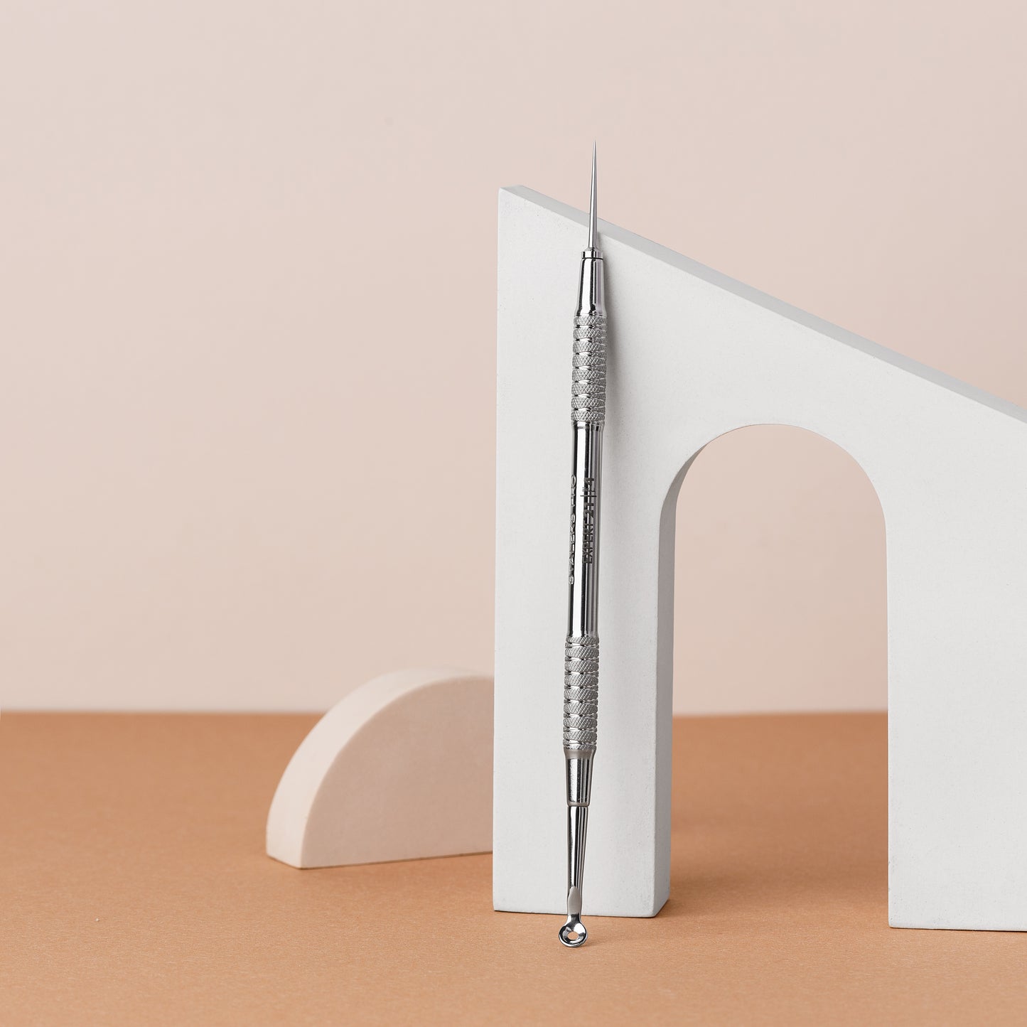 A STALEKS PRO Beauty care double-ended tool (UNO small and Vidal needle straight) EXPERT 21 TYPE 4 made of AISI 420 stainless steel propped vertically against a white arch-shaped cutout, all set against a soft peach-colored background.