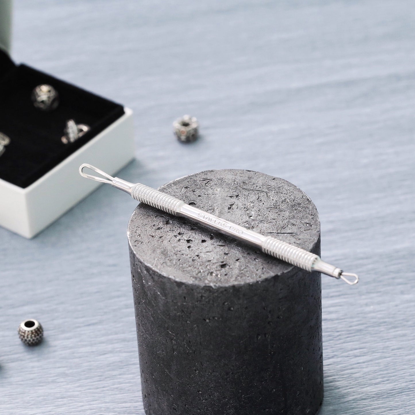 A close-up photo of a Staleks Blackhead Remover double-ended tool (2 loops) EXPERT 30 TYPE 1 stainless steel ear wax removal tool placed on a textured gray cylinder, with beads and a jewelry box blurred in the background.