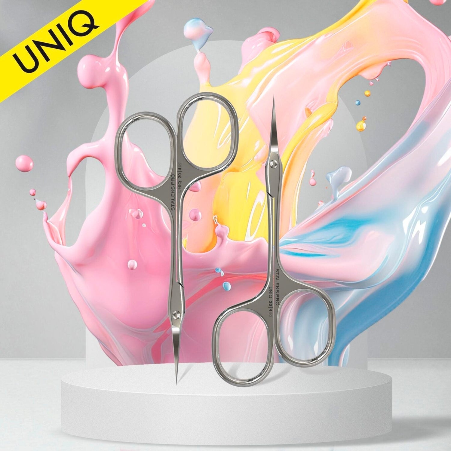 Two silver manicure scissors stand upright on a white platform, with colorful splashes of pink, yellow, and blue paint swirling in the background. A yellow "UNIQ" banner appears in the top left corner.