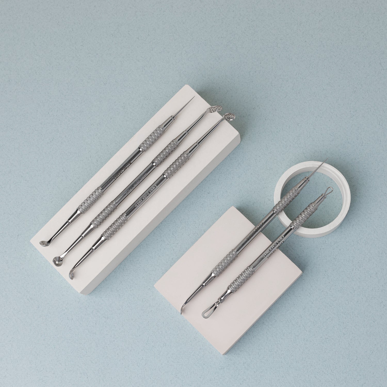 A set of stainless steel skincare tools with textured handles is neatly arranged on two white rectangular blocks. The tools include various types of extractors and tweezers, designed for facial care, displayed on a light blue background.