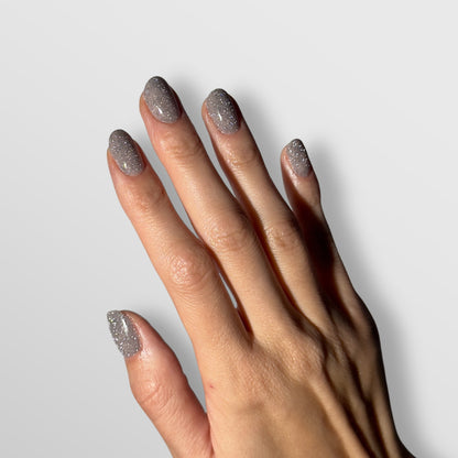 A hand with neatly manicured nails painted in Lumore’s LUMORE PARIS Reflective Gel Polish in the shade Margarita (9 ml), a sparkly silver hue, is shown against a plain light gray background.