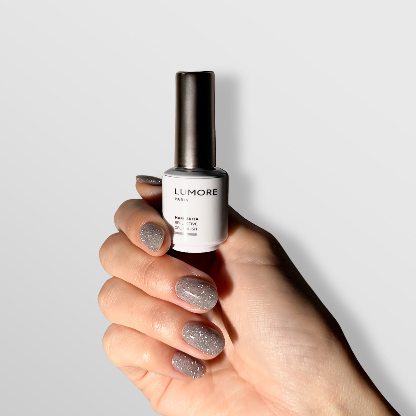 A hand with glittery gray reflective gel polish holds a white bottle of LUMORE PARIS Reflective Gel Polish, Margarita 9 ml by Lumore against a light gray background.