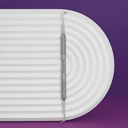 The STALEKS PRO Pedicure Curette EXPERT 20 TYPE 2 (Double-Ended Curette) by Staleks, featuring textured grips, rests horizontally on a white ridged oval platform against a purple background—a versatile tool for ingrown nail care.