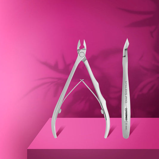 Two silver manicure tools—Staleks PRO Professional Cuticle Nippers EXPERT 20 8mm and a cuticle pusher—stand upright on a pink surface with leafy shadows against a pink background.