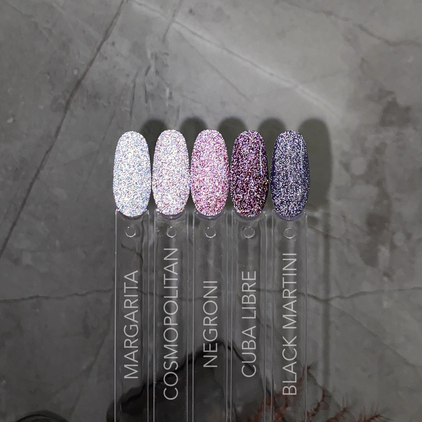 Five shimmering nail swatches painted with Lumore’s LUMORE PARIS Reflective Gel Polish in shades ranging from sparkling white to deep purple—named Margarita, Cosmopolitan, Negroni, Cuba Libre, and Black Martini—are arranged on a gray marble background, ideal for displaying dazzling nail art created with this reflective gel polish.