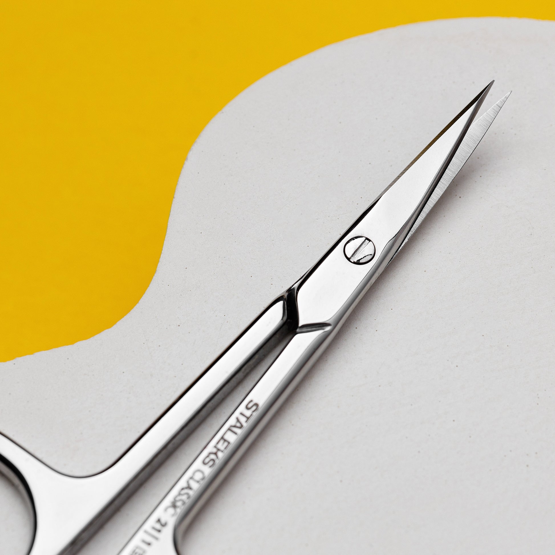 The STALEKS PRO Cuticle Scissors CLASSIC 21 TYPE 1 by Staleks, made of sharp stainless steel, rest on a white curved surface with a bright yellow background—ideal for precise manicures and pedicures.