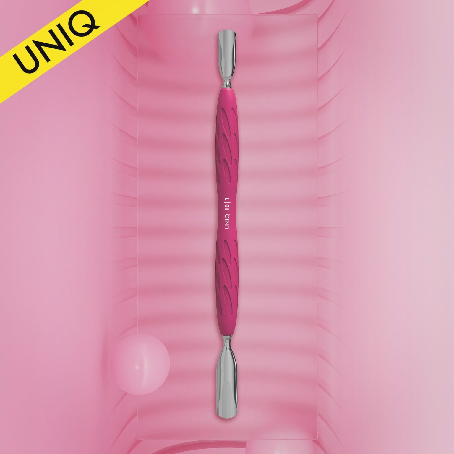 A pink dual-ended STALEKS PRO Gummy Manicure pusher with a silicone handle, featuring both wide and narrow rounded pushers, is displayed vertically on a textured pink background. In the top left, a yellow corner flag reads "UNIQ." Two soft pink round objects in the background enhance its appeal.