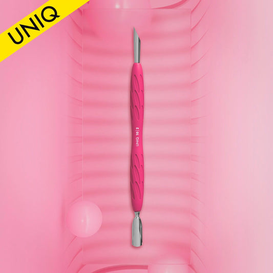A pink STALEKS PRO Gummy Manicure Pusher with a silicone handle and textured grip stands vertically on a ribbed surface. This tool, perfect for manicure and pedicure tasks, features metallic tips—a narrow rounded pusher on one end and a slanted pusher on the other. A yellow corner label displays "UNIQ," while two pink spheres are positioned at the bottom.