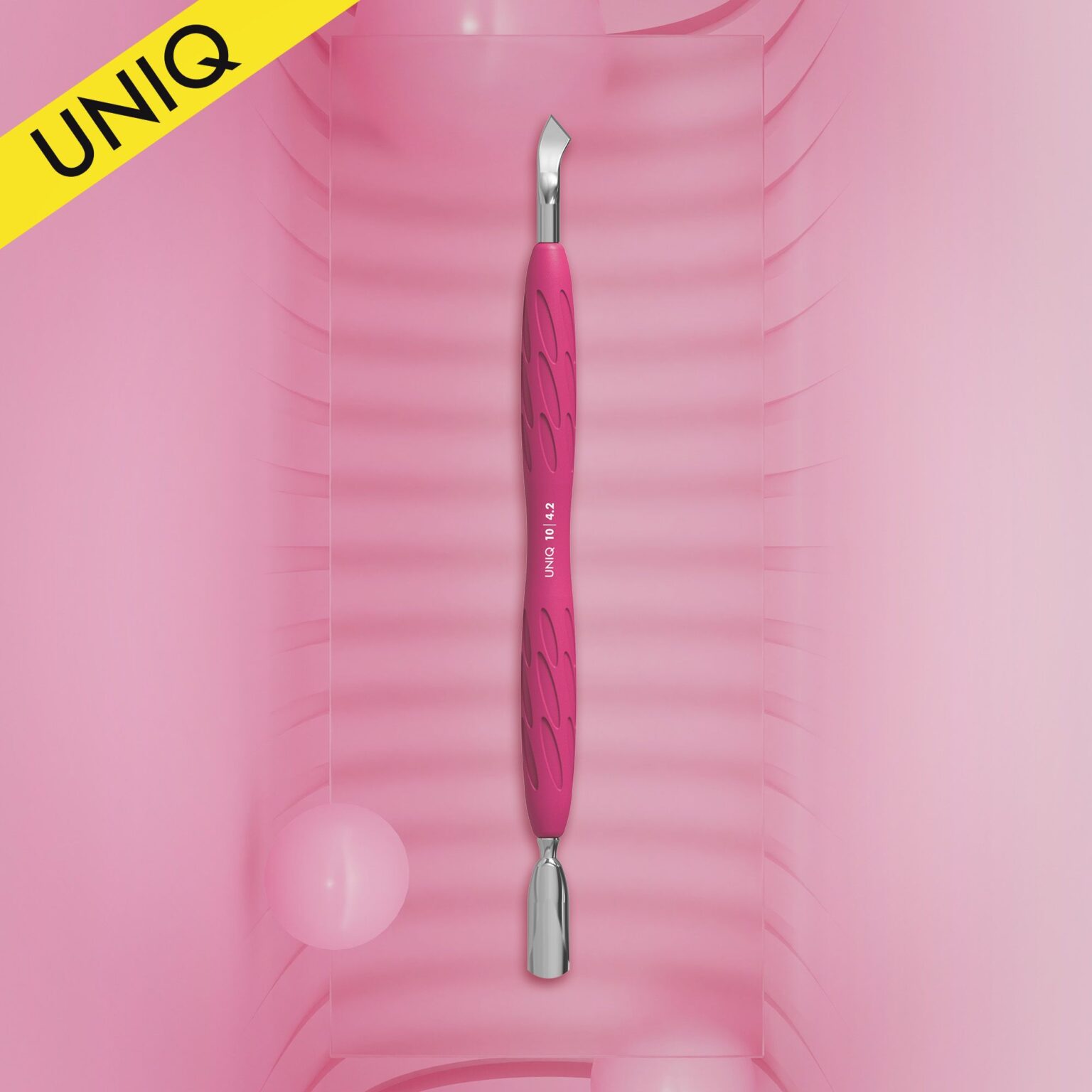 The STALEKS PRO Gummy Manicure Pusher with Silicone Handle UNIQ 10 TYPE 4.2 by Staleks, featuring a narrow rounded pusher and bent blade, stands vertically on a textured pink surface with rounded edges. A yellow corner labeled "UNIQ" is at the top left, accompanied by two pink spheres.