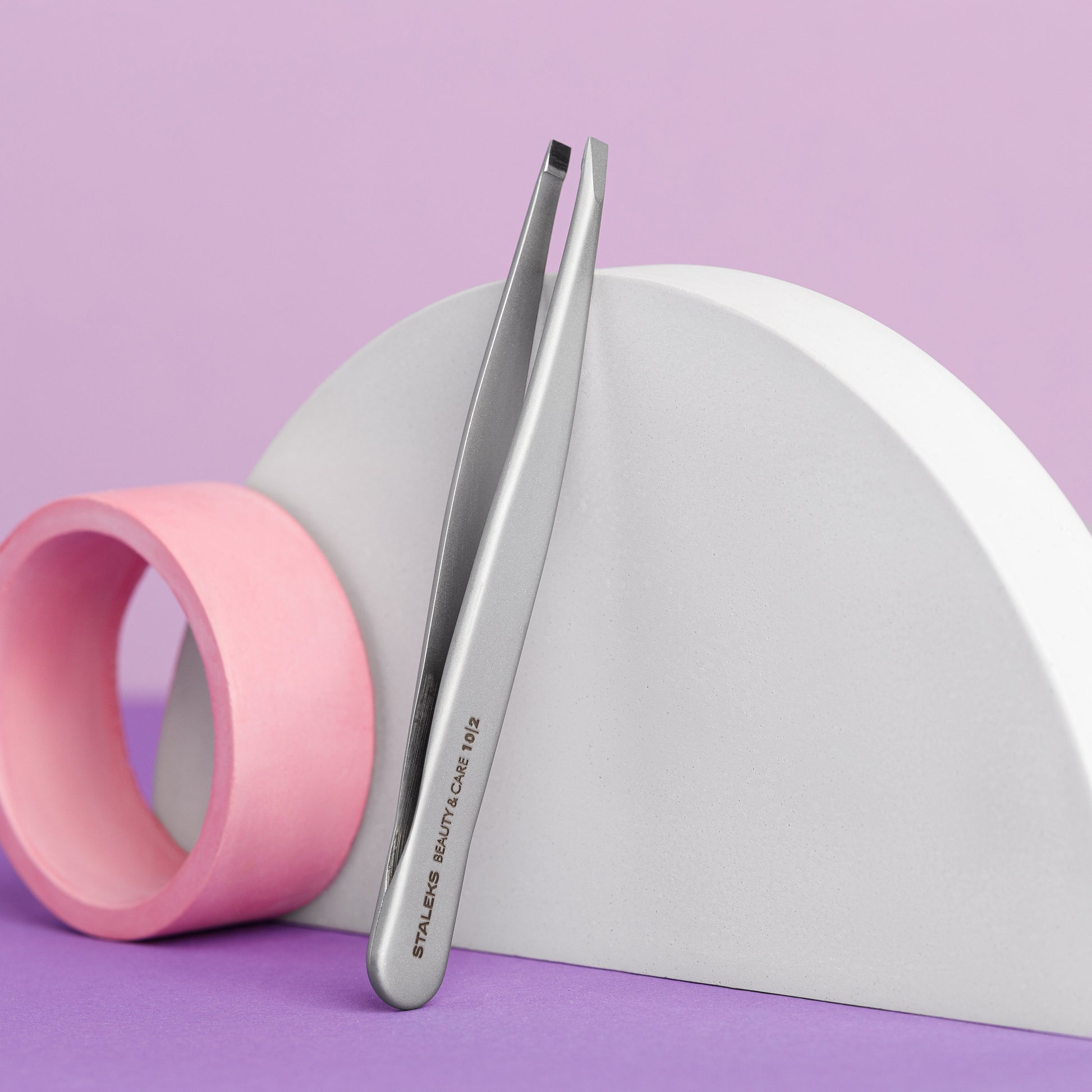 A pair of STALEKS PRO Eyebrow Tweezers Beauty & Care 10 Type 2 by Staleks leans on a white semicircular prop beside a pink cylinder, all on a purple background—ideal for shaping thick eyebrows.