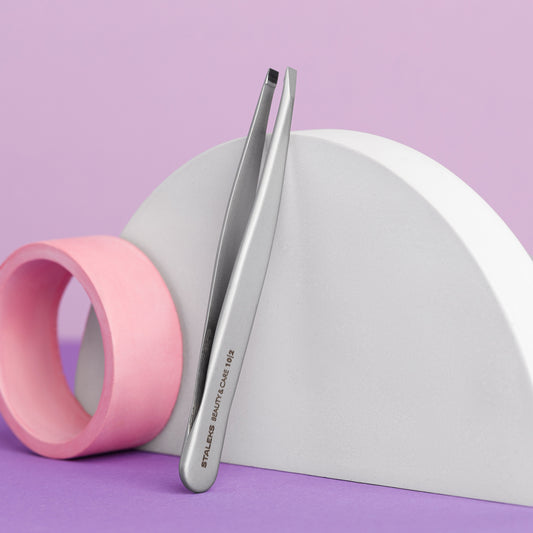 A pair of STALEKS PRO Eyebrow Tweezers Beauty & Care 10 Type 2 by Staleks leans on a white semicircular prop beside a pink cylinder, all on a purple background—ideal for shaping thick eyebrows.