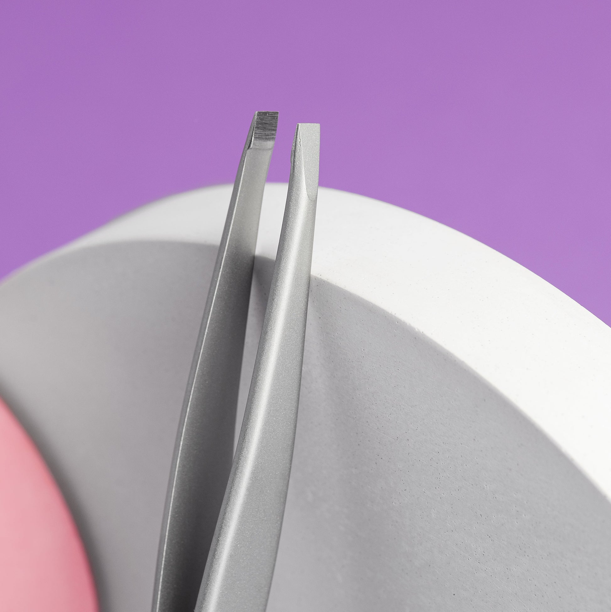 A close-up of two Staleks STALEKS PRO Eyebrow Tweezers Beauty & Care 10 Type 2 with angled tips resting on a curved white surface against a purple background.