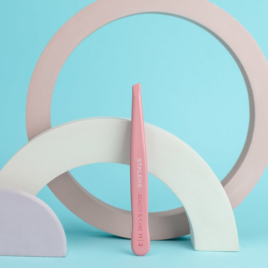A pink STALEKS PRO Eyebrow Tweezer BEAUTY & CARE 11 TYPE 3 (wide beveled) stands upright among pastel arches and a ring, crafted from stainless steel AISI 420, against a light blue background.