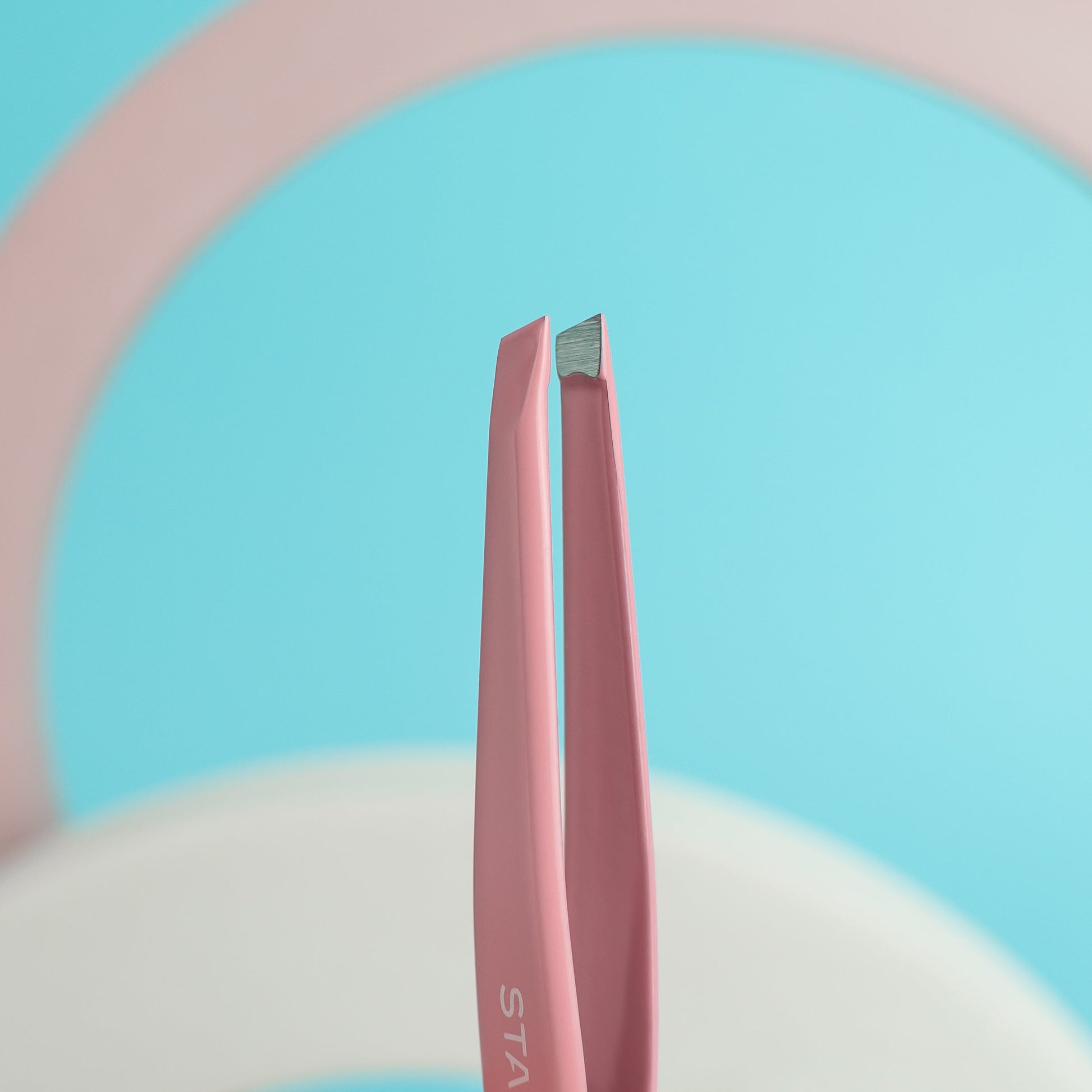 A close-up of the Staleks STALEKS PRO Eyebrow Tweezers BEAUTY & CARE 11 TYPE 3 (wide beveled) in pink, held upright against a pastel blue and pink background. Made from durable stainless steel AISI 420 for long-lasting precision.