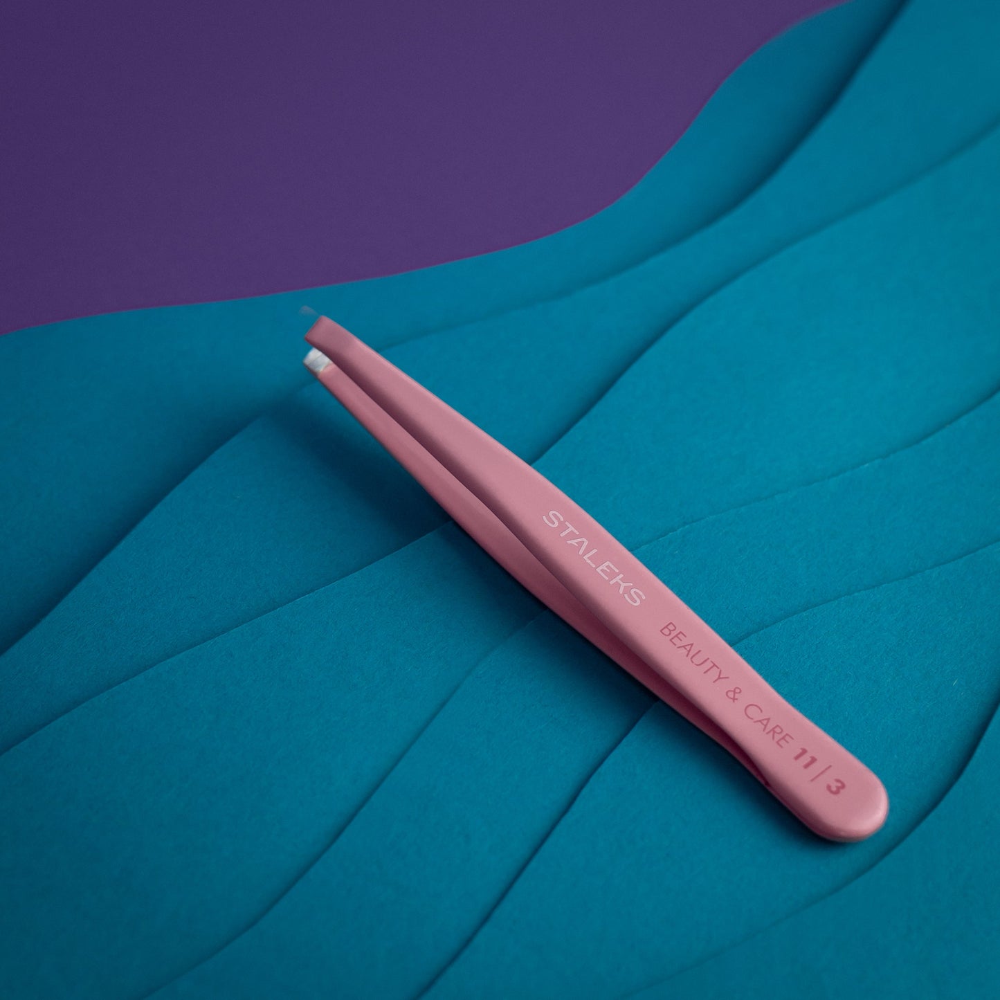 The pink STALEKS PRO Eyebrow Tweezers BEAUTY & CARE 11 TYPE 3 (wide beveled), made from AISI 420 stainless steel, are shown on a textured blue surface with a purple background in the corner.