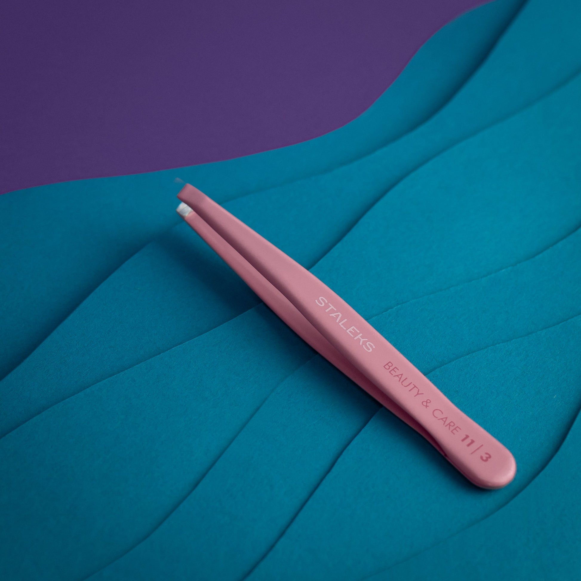 The pink STALEKS PRO Eyebrow Tweezers BEAUTY & CARE 11 TYPE 3 (wide beveled), made from AISI 420 stainless steel, are shown on a textured blue surface with a purple background in the corner.