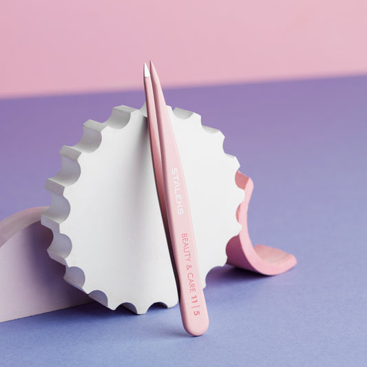 A pair of pink STALEKS PRO Eyebrow Tweezers BEAUTY & CARE 11 TYPE 5 (Point) by Staleks rests against a white, gear-shaped object on a purple and pink background.