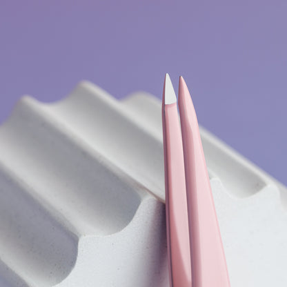 A pair of Staleks STALEKS PRO Eyebrow Tweezers BEAUTY & CARE 11 TYPE 5 (Point), in pink stainless steel with white pointed tips, rests on a textured white surface against a purple background.